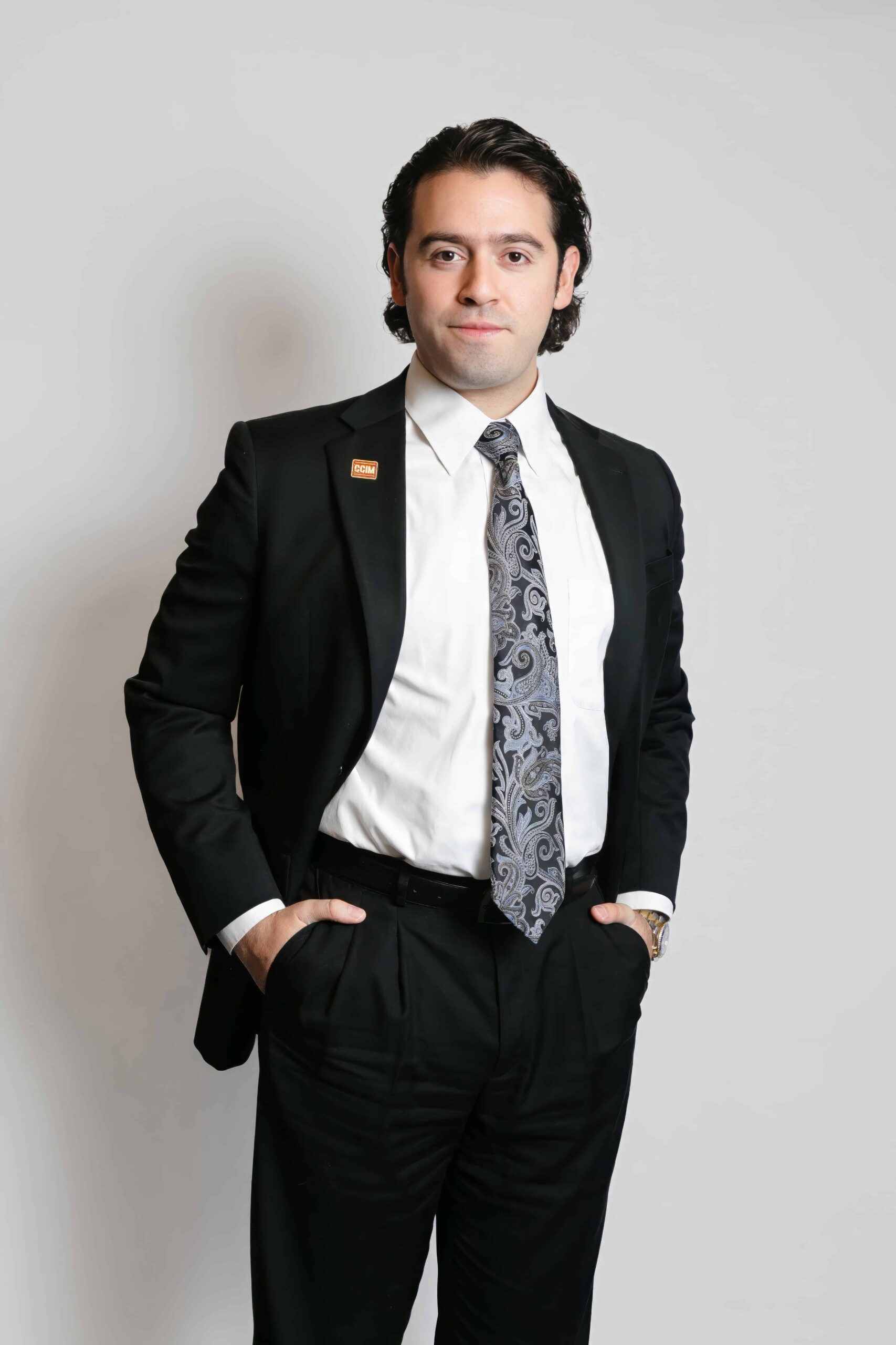 Man in a black suit with a patterned tie stands against a plain white background with hands in his pockets, portraying the professionalism of El Paso commercial real estate brokerage.